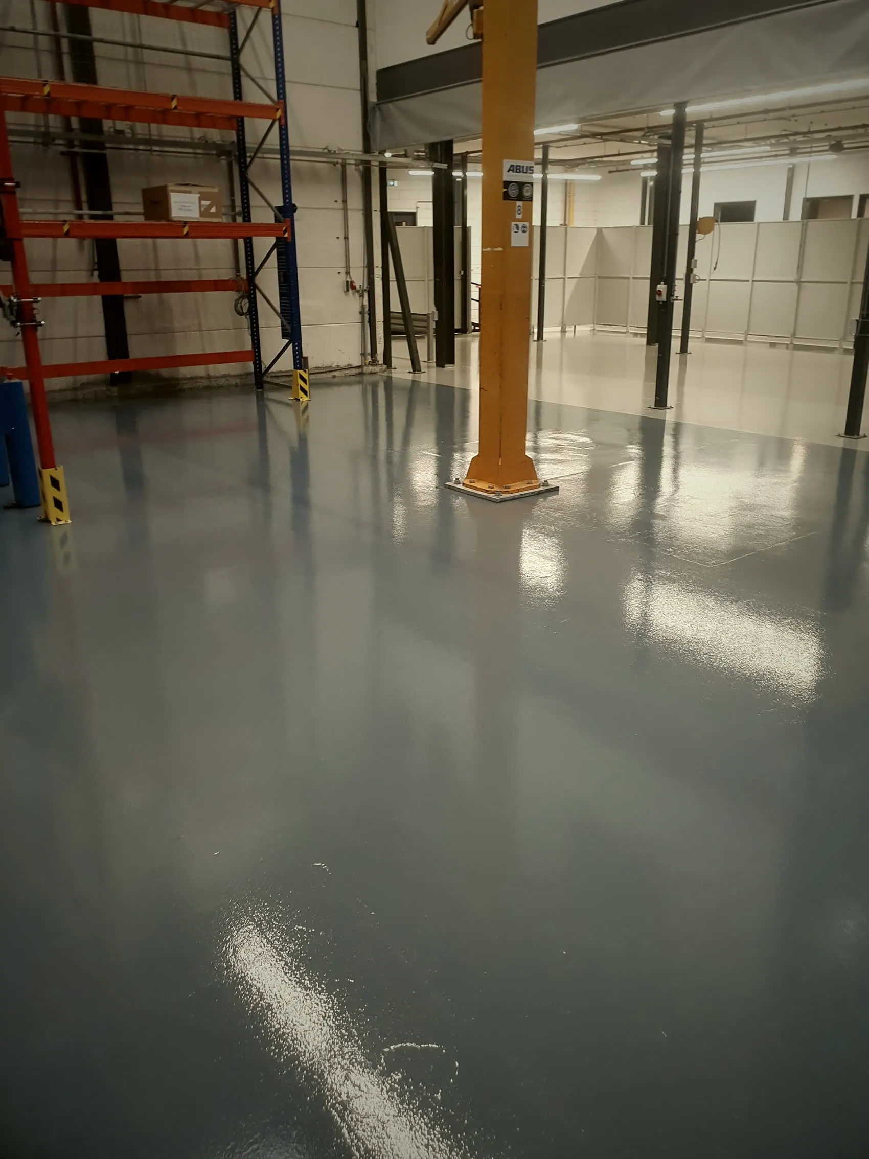 Magazijn met grijze epoxyvloer die de daklichten weerspiegelt, met stellingen en kolommen